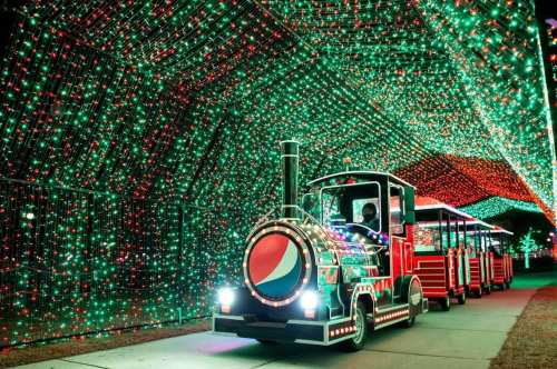 A colorful train decorated with lights travels through a tunnel of vibrant red and green holiday lights.