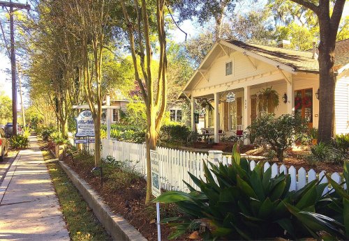 A charming white house with a porch, surrounded by greenery and a white picket fence, along a tree-lined sidewalk.