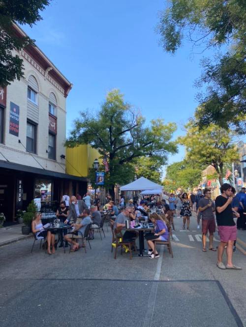 A bustling street scene with outdoor dining, people socializing, and tents set up under clear blue skies.