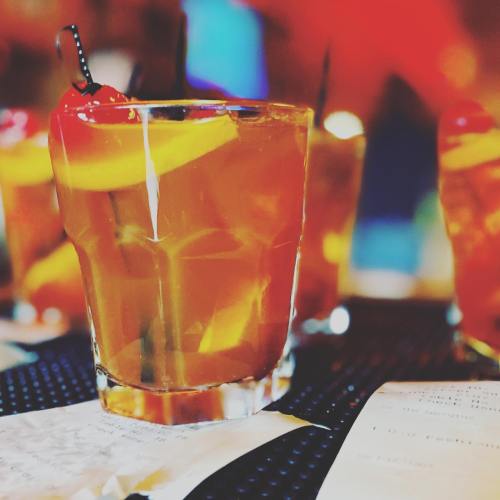 A close-up of a cocktail glass with a cherry and lemon slice, surrounded by other drinks and a menu on a table.