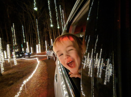 A joyful child leans out of a car window, surrounded by glowing holiday lights in a nighttime setting.