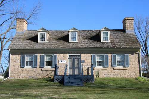 Historic stone house with a sign reading "OPEN," featuring multiple windows and a grassy lawn under a clear blue sky.