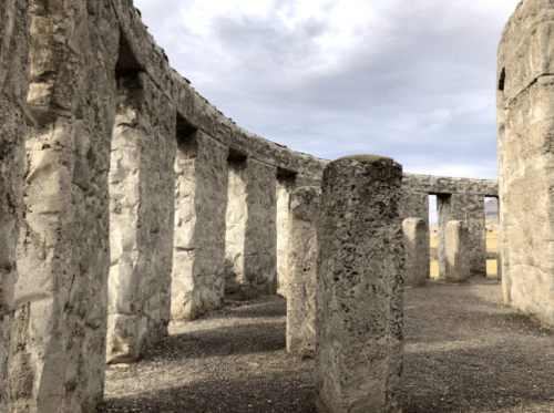 Ancient stone structure with tall, weathered pillars under a cloudy sky. Ground is gravelly and open.