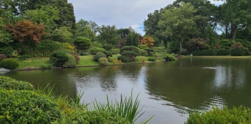 A serene garden with lush greenery and a calm pond, surrounded by trees and colorful shrubs under a cloudy sky.