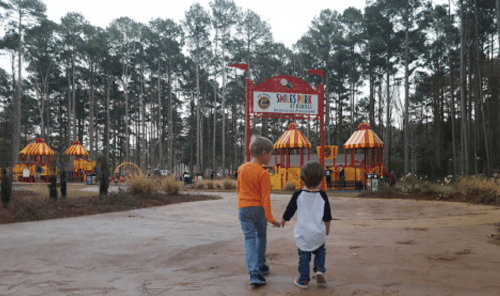 Two children walk hand in hand towards a colorful playground surrounded by tall trees.