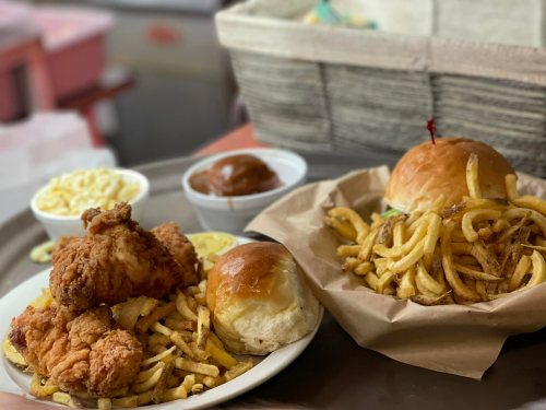 A plate of fried chicken with fries and rolls, alongside a burger with fries in a basket and a side of macaroni.