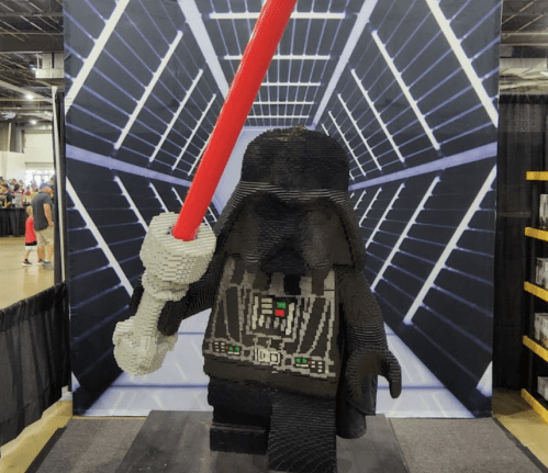 A large LEGO sculpture of a character holding a red lightsaber, set against a space-themed backdrop.