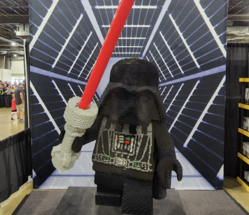 A large LEGO sculpture of a character holding a red lightsaber, set against a space-themed backdrop.