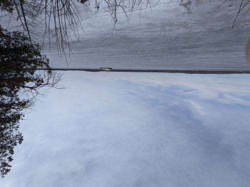 A snowy landscape with a frozen body of water and a distant truck on a road under a cloudy sky.