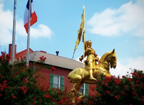 A golden statue of a knight on horseback, holding a flag, surrounded by red flowers and flags in the background.