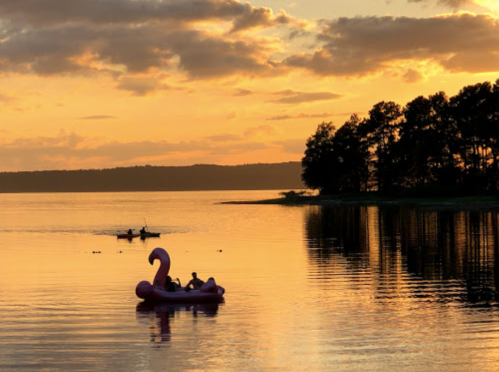 A pink flamingo float with two people on a calm lake at sunset, with a kayak in the background and trees along the shore.