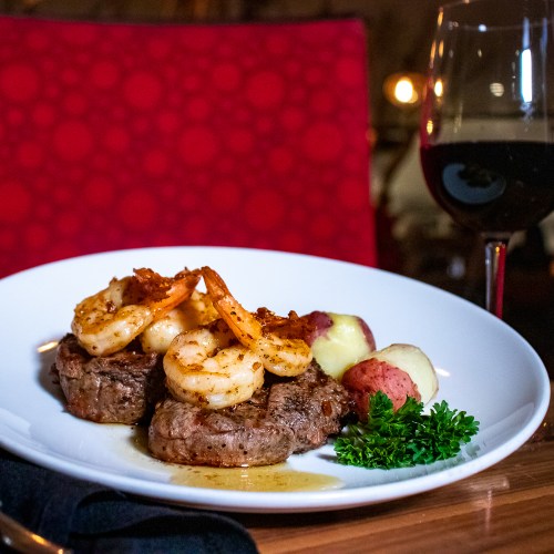 A plate featuring steak topped with shrimp, served with potatoes and parsley, alongside a glass of red wine.