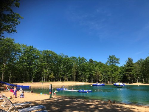 A serene lake surrounded by trees, with people enjoying the beach and inflatable boats on the water under a clear blue sky.