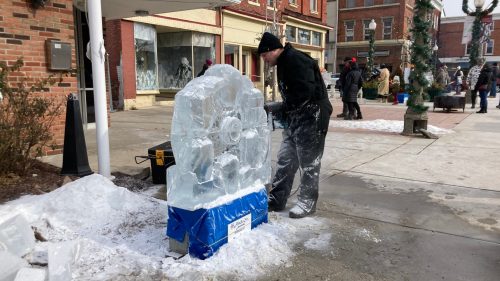 A person carves an ice sculpture in a snowy outdoor setting, with buildings and a crowd in the background.