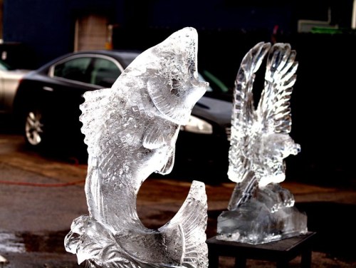 Two ice sculptures: one of a fish and another of an eagle, displayed outdoors with a car in the background.