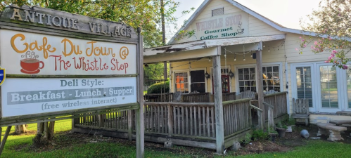 A quaint coffee shop with a sign reading "Café Du Jour @ The Whistle Stop," surrounded by greenery.