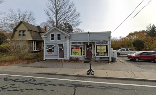 A small blue building with signs advertising food, next to a house, along a road with parked cars and autumn trees.