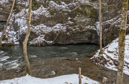 A serene river flows through a snowy landscape, surrounded by rocky cliffs and bare trees.