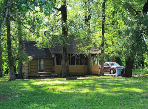 A rustic cabin surrounded by trees, with a picnic table in front and a car parked nearby on a grassy area.