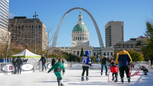 Skaters enjoy an outdoor rink with the Gateway Arch and historic buildings in the background on a sunny day.