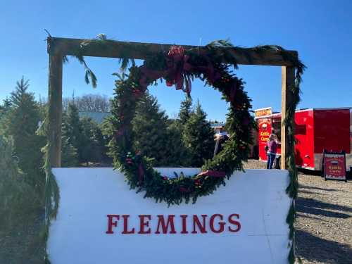 A festive wreath hangs on a wooden frame with "Flemings" written below, surrounded by Christmas trees and a clear blue sky.