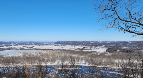 A panoramic winter landscape featuring snow-covered fields and hills under a clear blue sky.
