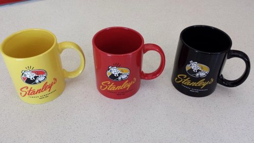 Three mugs in yellow, red, and black, each featuring the logo "Stanley's" on a light-colored surface.