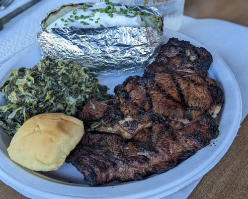 A plate featuring grilled meat, collard greens, a baked potato wrapped in foil, and a roll.
