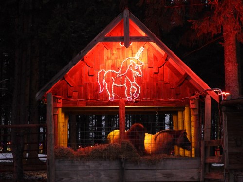 A wooden barn illuminated with red lights, featuring a unicorn neon sign and two horses resting on hay inside.