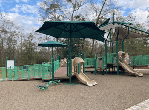 Playground with two slides, shaded areas, and a climbing structure, surrounded by trees and blue sky.