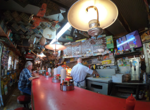 A diner interior with a red counter, two patrons, and a cook preparing food amidst colorful vintage decor.