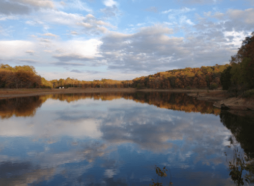 A serene lake reflecting autumn trees and a cloudy sky, surrounded by colorful foliage.
