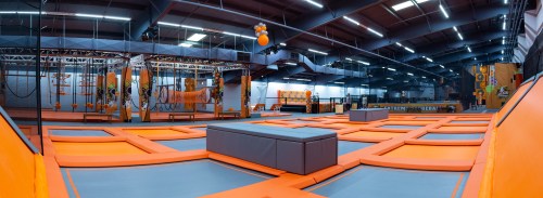 A spacious indoor trampoline park with orange and gray trampolines, climbing structures, and safety nets.
