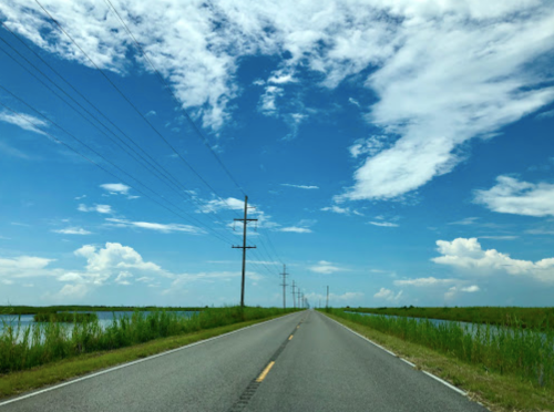 A straight road lined with power poles, flanked by water and lush greenery under a blue sky with scattered clouds.