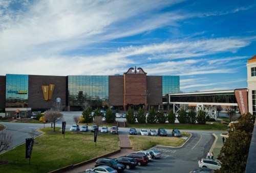 A modern building with large glass windows and a brick facade, surrounded by greenery and parked cars under a blue sky.