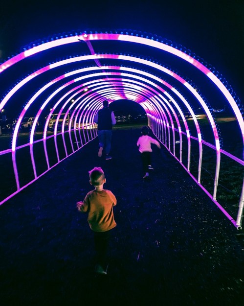 Two children run through a brightly lit, colorful tunnel at night, with an adult walking ahead of them.