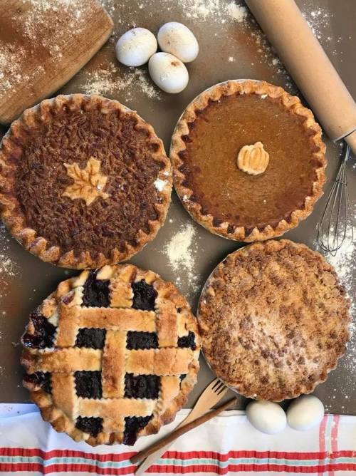 Four delicious pies on a floured surface, including pecan, pumpkin, berry, and crumb-topped, with eggs and a rolling pin.