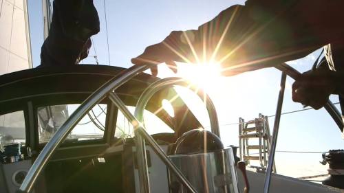 A person steering a sailboat with the sun shining brightly in the background, creating a lens flare effect.