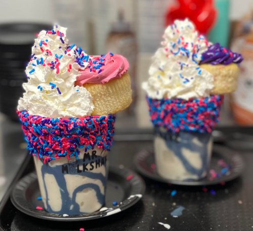 Two colorful milkshakes topped with whipped cream, sprinkles, and a cookie, served in cups on a black tray.