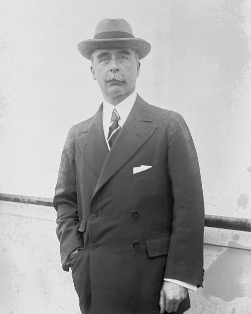 A man in a formal suit and hat stands confidently, with a mustache and a pocket square, against a plain background.