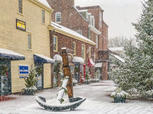 Snow-covered street scene with shops and a decorative anchor in the foreground, creating a wintery atmosphere.