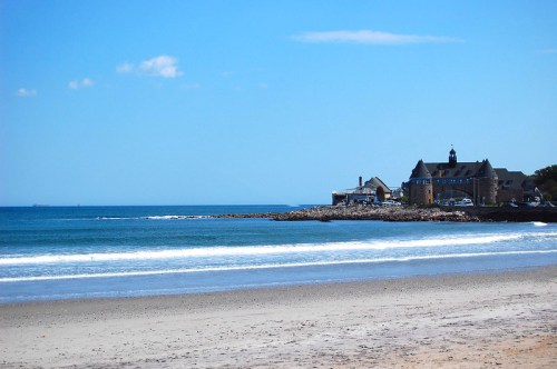 A serene beach scene with gentle waves, a sandy shore, and a coastal building under a clear blue sky.