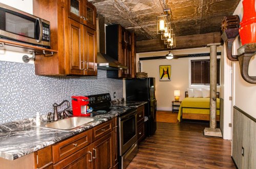 Modern kitchen with dark wood cabinets, stainless appliances, and a cozy bedroom in the background featuring a yellow bed.