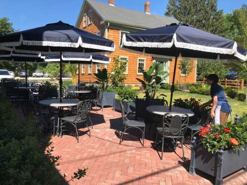 Outdoor dining area with tables and umbrellas, surrounded by greenery and flowers, near a wooden house.