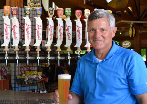 A smiling man in a blue shirt holds a beer in front of a row of beer taps at a bar.