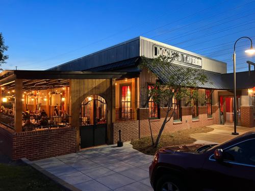 A cozy restaurant exterior at dusk, featuring outdoor seating and warm lighting, with a brick facade and large windows.