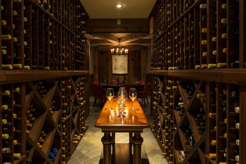 A cozy wine cellar with wooden racks filled with bottles, a table with glasses, and a dining area in the background.