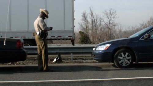 A police officer stands on the highway, checking his phone near two parked cars. Trees are visible in the background.