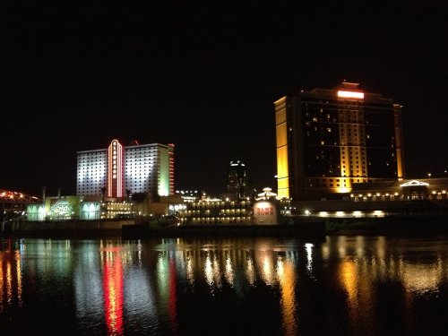 Nighttime view of illuminated buildings along a river, reflecting colorful lights on the water's surface.