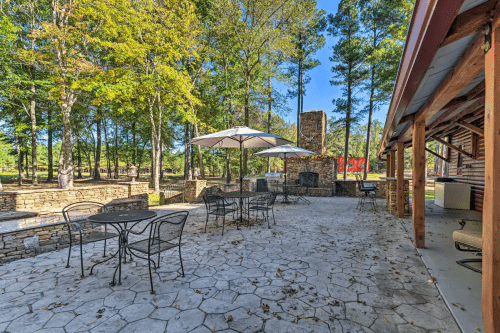 Outdoor patio area with stone flooring, tables, umbrellas, and a fireplace surrounded by trees.